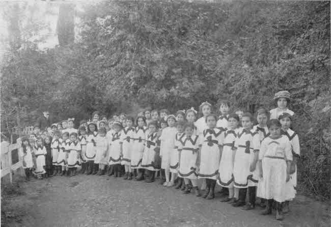 The girls are arranged standing
generally in three tiers arranged in a slight semicircle receding to the left.
The nearer and somewhat older-appearing girls are to the right and closer to
the camera. The girls are all wearing identical white dresses with trim.
Most do not have head coverings, but a few do. The girls appear to mostly be
of pre-teen age. There are approximately fifty girls plus two adults,
one male and one female, with one adult near each end of the semicircle.