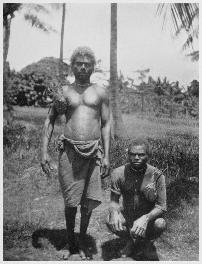 Two native men, one standing on the left, the other squatting on the right, facing the viewer.